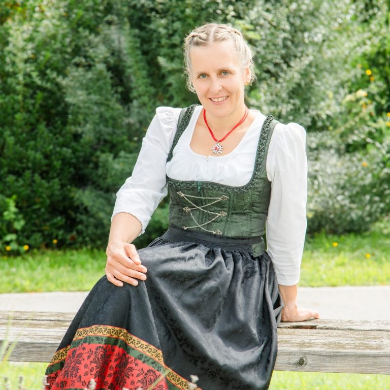 Eva Herz Frau im traditionellen Dirndl mit grünem Mieder und schwarzer Schürze, lächelnd.