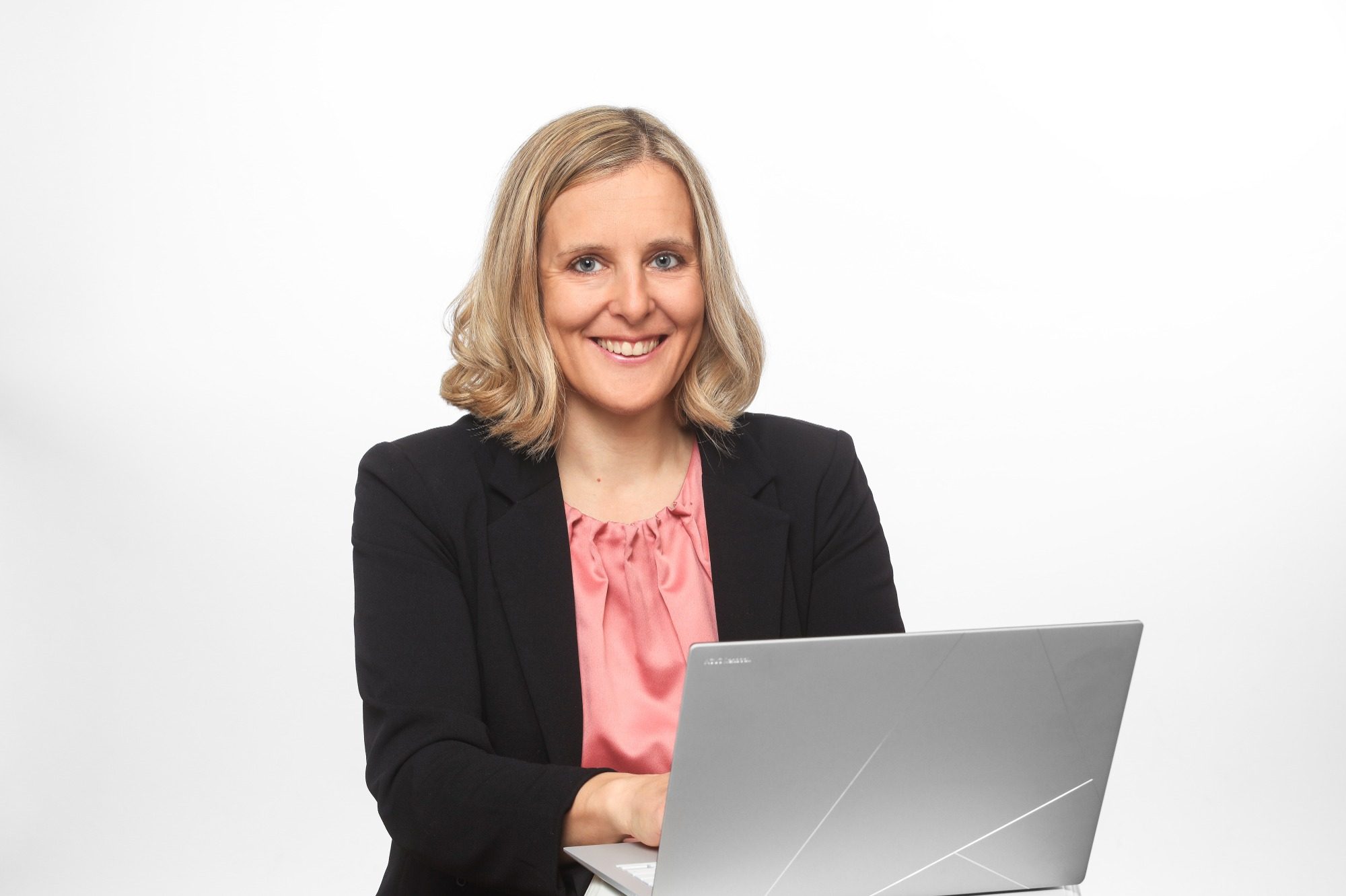 Frau mit blonden, lockigen Haaren sitzt lächelnd am Laptop.
