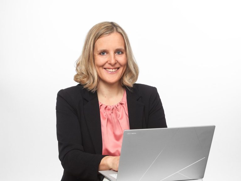 Eva Herz Lächelnde Frau mit blonden Locken, die an einem Laptop arbeitet.