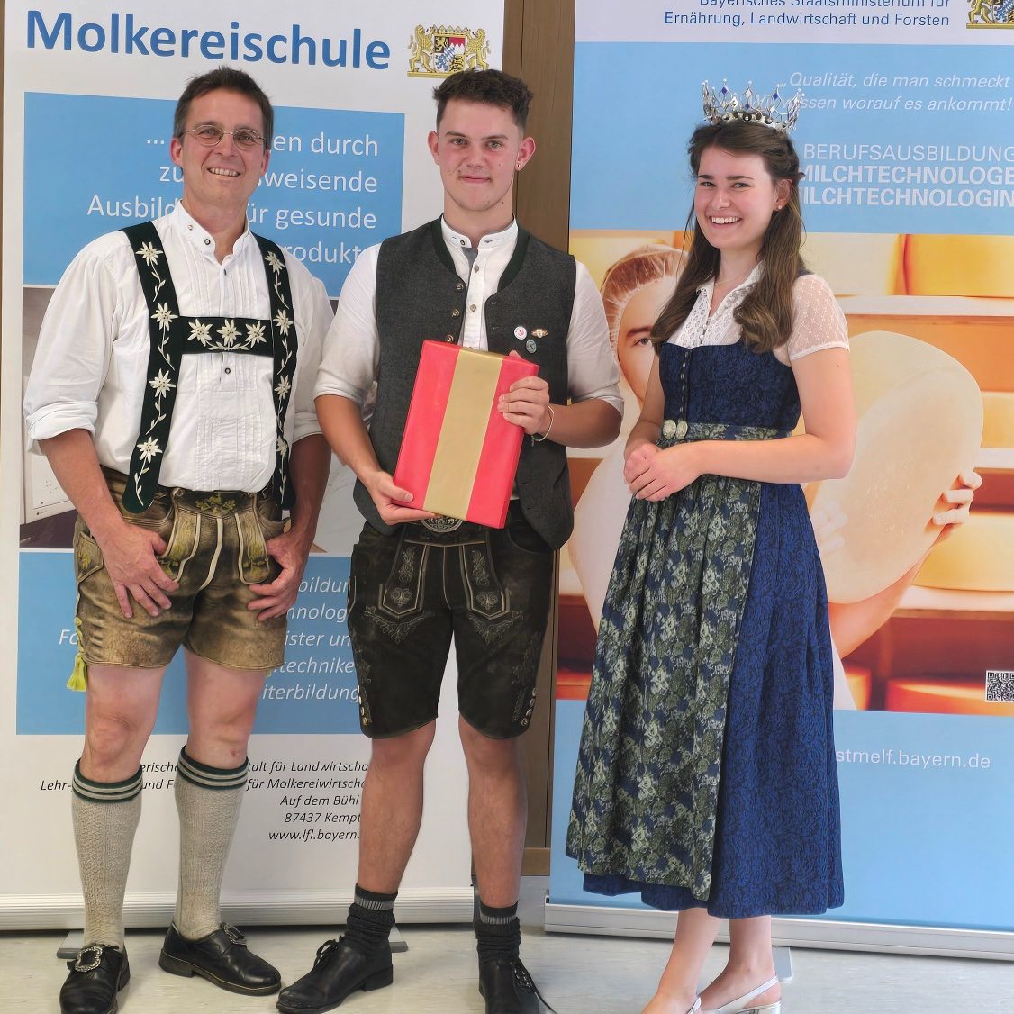 Ausbildung Drei Personen in traditioneller Kleidung stehen vor einem Banner der Molkereischule.