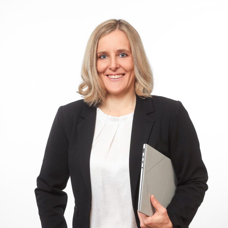 Eva Herz Frau mit blonden Haaren, lächelnd, in einem schwarzen Blazer, hält einen Laptop.