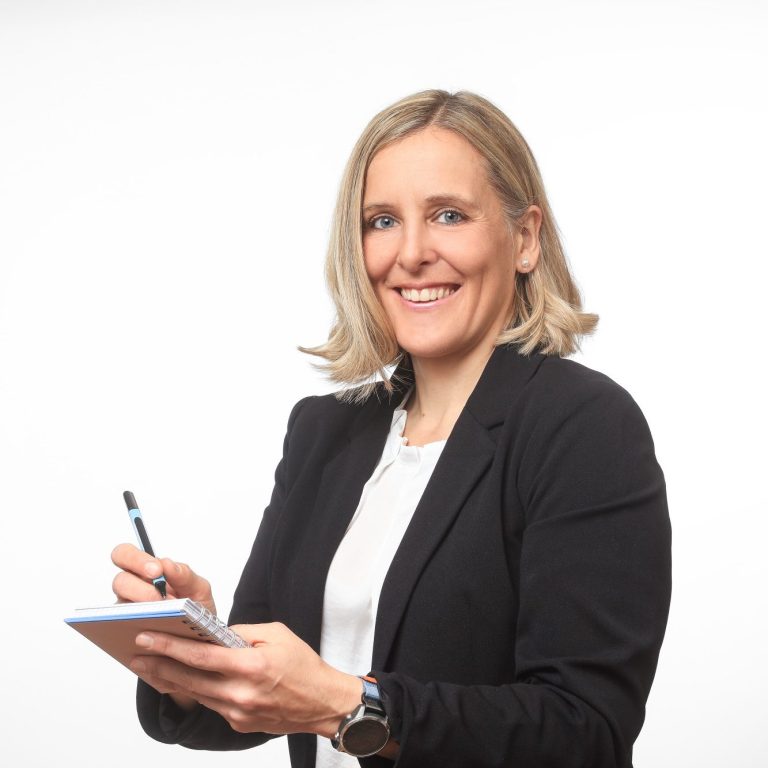 Text & Storytelling Frau in schwarzer Blazer-Jacke hält Notizblock und Kugelschreiber, lächelt freundlich.